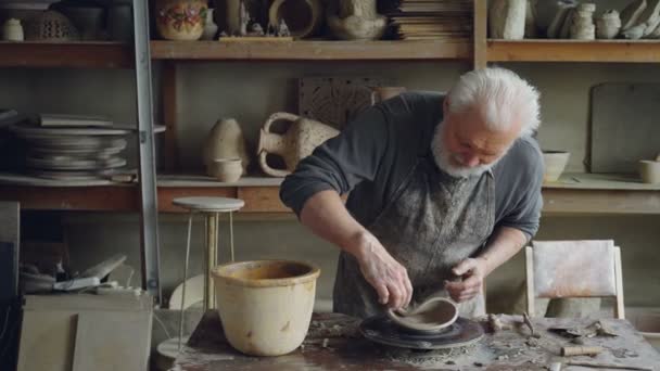 Kreativa skulptör gör ovanligt bred skål stående på arbetsbordet i hans verkstad. Skäggig senior man är liiking på sin clayware samt utvecklar design och kvalitet. — Stockvideo