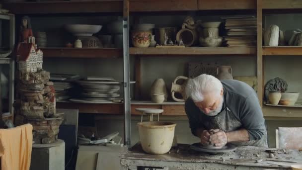 経験豊富な彫刻家上級大人は、ホイールを投げに粘土団子から彫刻を作っています。彼はそれと泥だらけの手で触れることを柔らかくために粘土に水をまいてください。. — ストック動画