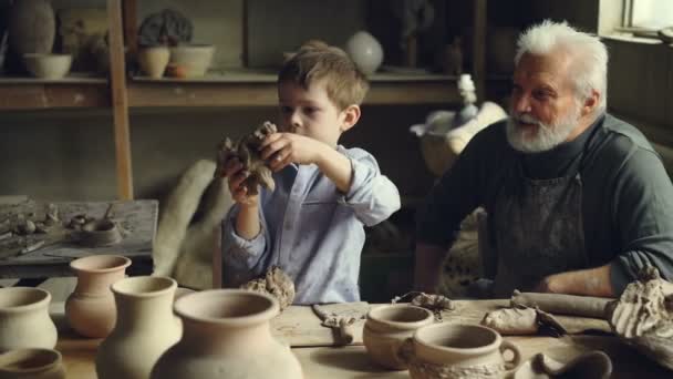 Profesyonel yaşlı potter torunu kil çömlek stüdyosunda oynamaktan izliyor. Yaratıcı çocuk çamurlu giysili oyuncak kil yapmak ve bununla eğleniyor. — Stok video