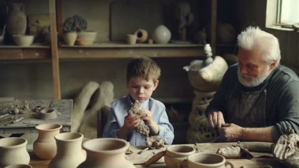 Професійний майстер і його маленький онук розмовляють і замішують глину на робочому столі в затишній студії. . — стокове відео