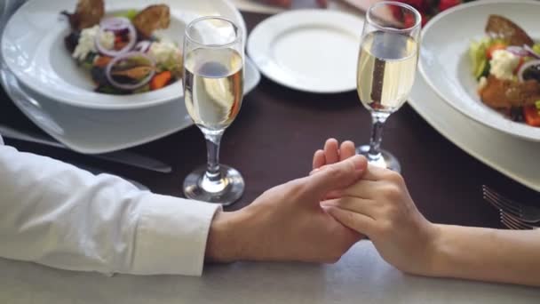 Gros plan de la main masculine tenant et caressant la main féminine sur la table avec des verres à champagne et des assiettes. Relation romantique, amour et concept gastronomique . — Video