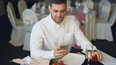 Sevgi dolu erkek mücevher kutusu açarak ve yüzüğe arayarak smartphone, kullanan onun kız restoranında beklemektedir. Buket gül ve şampanya görünür.