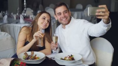 Çekici sevgi dolu çift selfie restoranda yemek yerken smartphone kullanarak şampanya bardağı ile alıyor. Onlar gülümseyen, öpüşme ve kameraya bakarak poz.