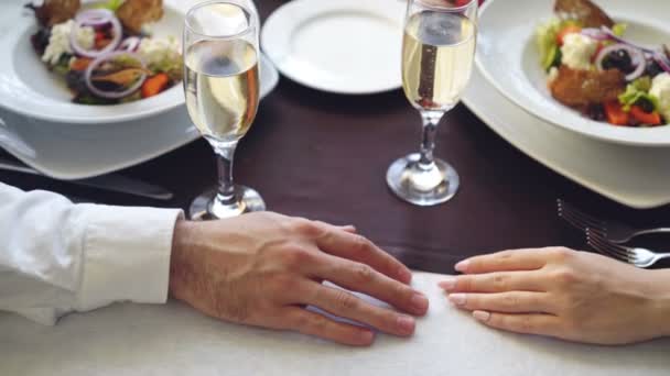 Close-up shot of young lovers touching and holding hands in classy restaurant. Table with sparkling champagne glasses, flatware and food in background. — Stock Video