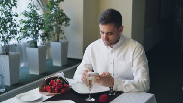 Joven enojado bien vestido está esperando a su novia en el restaurante, usando un teléfono inteligente, abriendo joyero y mirando el anillo y luego dejando, las flores dejadas en la mesa . — Vídeo de stock