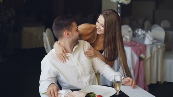Mujer joven y bonita está saludando a su novio en el restaurante cerrando los ojos con las manos, abrazándolo y besándolo, luego tomando flores y sentándose a la mesa delante de él . — Vídeos de Stock