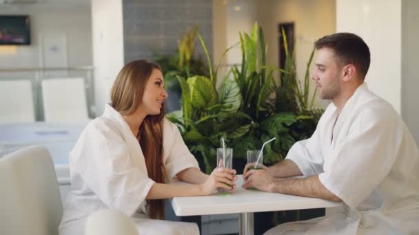 Jóvenes alegres están hablando, tintinear vasos de cóctel y beber mientras están sentados en la mesa en el spa de día. Cómodos muebles blancos y planta verde son visibles . — Vídeos de Stock