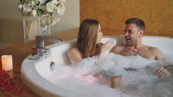 Vrij vrolijk meisje is blazen zeep schuim en plezier met haar bebaarde fience in bubbelbad in dag spa. Jongeren zijn lachen, genieten van weekend en praten. — Stockvideo