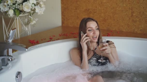 Mulher bonita em maiô está falando no telefone celular e bebendo champanhe enquanto toma banho no salão de spa. Comunicação, pessoas e conceito de relaxamento . — Vídeo de Stock