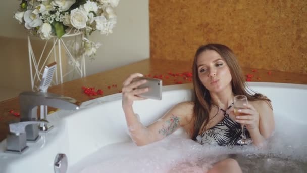 Красивая молодая женщина делает селфи с бокалом шампанского, используя смартфон в гидромассажной ванне в современном спа-салоне. Она улыбается и позирует, глядя в камеру . — стоковое видео