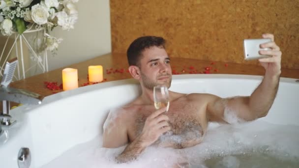 Guapo joven se está tomando selfie con copa de champán utilizando el teléfono inteligente en la bañera de hidromasaje en el moderno salón de spa. Él sonríe y posando mirando a la cámara . — Vídeos de Stock