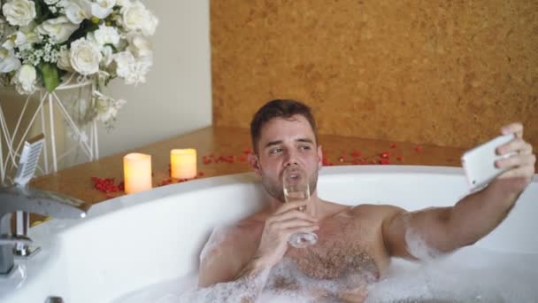 Young handsome man popular blogger is recording video in hot tub in day spa using smartphone. Burning candles, champagne glass and flowers are visible. — Stock Video