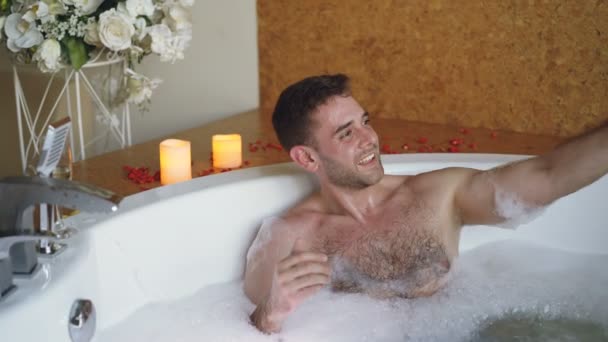 Joven barbudo famoso bloguero está grabando vídeo en la bañera de hidromasaje en el spa de día con el teléfono inteligente. Velas ardientes, copa de champán y flores son visibles . — Vídeos de Stock