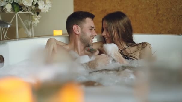 Menina bonita alegre está se divertindo em jacuzzi com seu namorado bonito reunindo espuma de sabão nas mãos e soprando-o. Conceito de relaxamento e bem-estar . — Vídeo de Stock