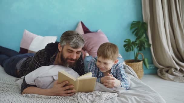 Pai carinhoso está lendo livro engraçado para seu filho, enquanto o menino está rindo e falando com seu pai. Cama de tamanho completo, almofadas brilhantes e plantas verdes são visíveis . — Vídeo de Stock