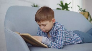 Sevimli küçük çocuk rahat kıyafetler içinde komik gülen ve yüksek sesle rahat koltukta yalan kitap okuyor. Mutlu çocukluk, okul öncesi eğitim ve hobi kavramı.