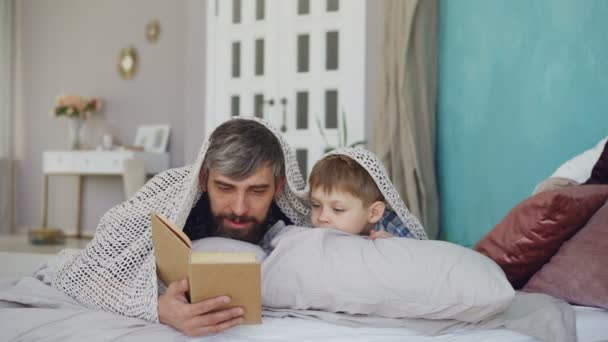 Padre cariñoso e hijo adorable están leyendo la historia de miedo y escondiéndose debajo de la manta mientras están acostados en la cama en casa. Feliz infancia, paternidad y amor concepto de la gente . — Vídeos de Stock