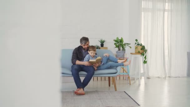 Skäggig man läser boken och visar illustrationer till sin son medan du kopplar av på soffan i ljus lägenhet. Enad familj, fritidsaktiviteter och höja barn koncept. — Stockvideo