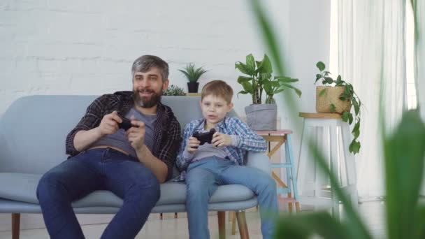 Baba ve genç çocuğunu evde kanepede video oyunu joystick düğmelere basarak ve konuşurken oynuyorlar. Mutlu bir aile ve modern teknoloji kavramı. — Stok video