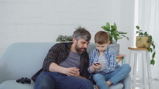 Der entzückende kleine Junge und sein fürsorglicher Vater bedienen sich des Smartphones, reden und lachen miteinander und zeigen auf den Bildschirm. Moderne Technologien und glückliches Familienkonzept. — Stockvideo