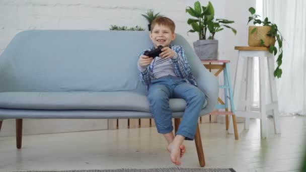 Joyeux petit enfant jouant au jeu vidéo seul s'amusant assis sur le canapé à la maison. Technologies modernes, enfance heureuse et concept de temps libre . — Video