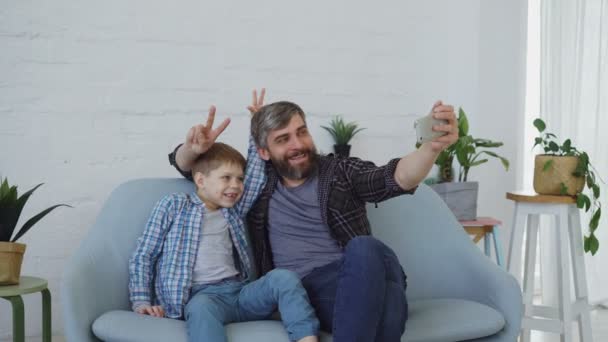 Cheerful handsome man is taking selfie with his cute little son using smartphone, they are posing, talking and gesturing. Self-portrait and happy family concept. — Stock Video