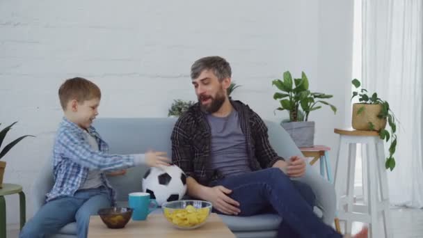 Vrolijke liefhebbende vader speelt met zijn zoontje gooien en schoppen Voetbal en eten snacks in lichte appartement. Gelukkige familie en sport concept. — Stockvideo