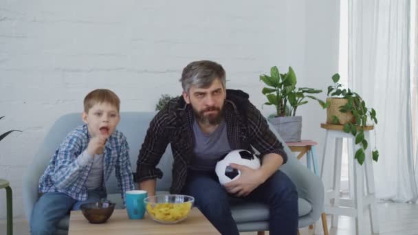 응원, 파이브와 함께 승리를 축 하 하 고 간식 먹는 집중된 아버지와 작은 아들 집에서 Tv에서 경기 하는 축구를 보고 있다. 행복 한 가족 및 스포츠 개념. — 비디오
