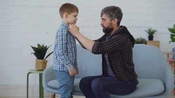 El padre cuidadoso está ayudando a su pequeño hijo a vestirse, abotonándose la camisa y alisándose el pelo. Padres cariñosos, infancia feliz y concepto de atención paterna . — Vídeos de Stock