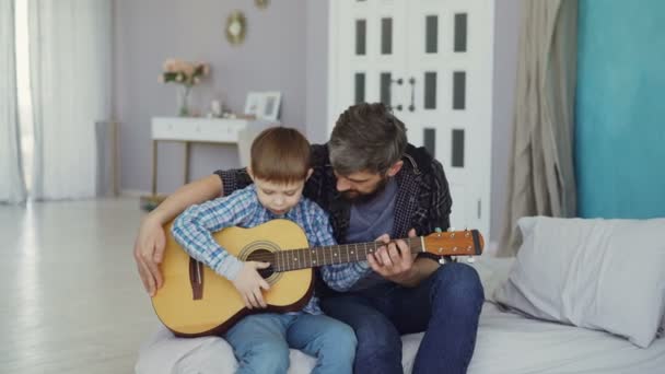 Junger Mann, der seinem talentierten kleinen Sohn das Gitarrespielen beibringt, während er im Bett in einer hellen Wohnung sitzt. Glückliche Kindheit, väterliche Liebe und Musikkonzept. — Stockvideo