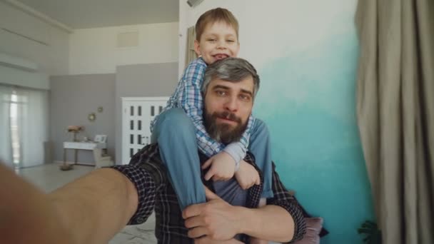 Punto de vista plano del hombre barbudo y su hijo sentado sobre sus hombros grabando video selfie, hablando y riendo en casa. Familia feliz y concepto de tecnología moderna . — Vídeo de stock