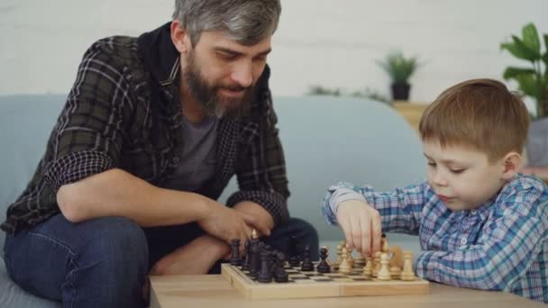 Inteligente niño pequeño está jugando ajedrez con su padre cariñoso tocando piezas de ajedrez y ajedrecistas en movimiento. Juego intelectual, familia feliz y concepto de generaciones . — Vídeo de stock