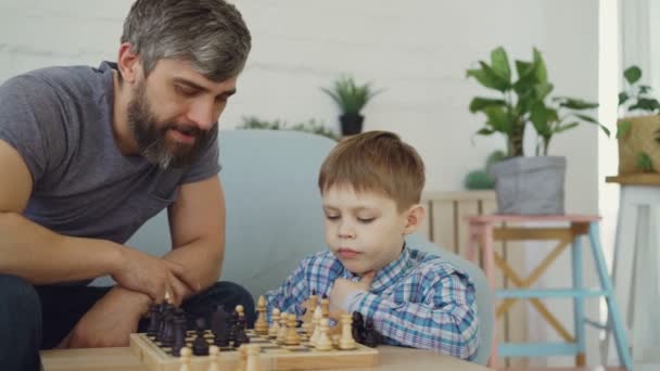 Серйозна дошкільна дитина грає в шахи зі своїм батьком, думаючи про наступний хід і переміщення шахових фігур, в той час як його батько вчить його тактики гри . — стокове відео