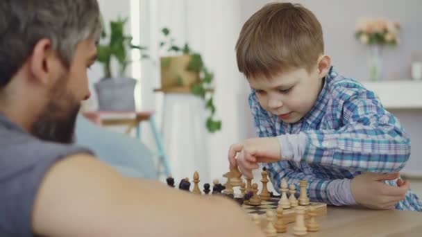 Умный мальчик учится играть в шахматы, двигая шахматы на борту и наслаждаясь игрой со своим отцом. Его отцы бородатые на переднем плане. . — стоковое видео