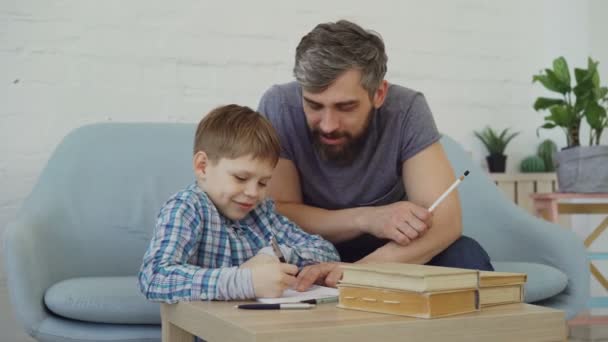 Niño feliz asiduo colegial está haciendo la tarea y hablar con su padre, niño pequeño está aprendiendo y papá está enseñando. Escuela junior y concepto de familia unida . — Vídeo de stock