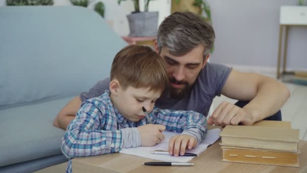 Schattig kind schrijft in notitieblok met zijn vader in de buurt van de tafel zitten en kijken naar zijn zoon. Voorschoolse educatie, Verenigde familie, vaderschap en jeugd concept. — Stockvideo