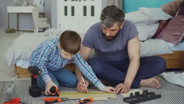 Батько і син вимірюють дерев'яний шматок, готуючись побудувати щось разом всередині будинку. Видимі різні інструменти та сучасні меблі . — стокове відео