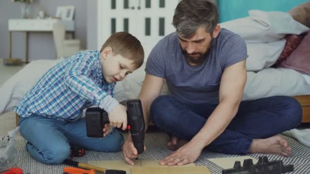 El niño está aprendiendo a usar un destornillador eléctrico mientras su padre explica cómo trabajar con una pistola de tornillo y fijar el tornillo en un pedazo de madera. Construcción y concepto familiar . — Vídeos de Stock