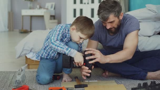 Padre attento sta insegnando al figlio a lavorare con cacciavite elettrico mentre il figlio sta cercando di utilizzare pistola a vite e fissare vite in pezzi di legno. Costruzione e concetto di famiglia . — Video Stock