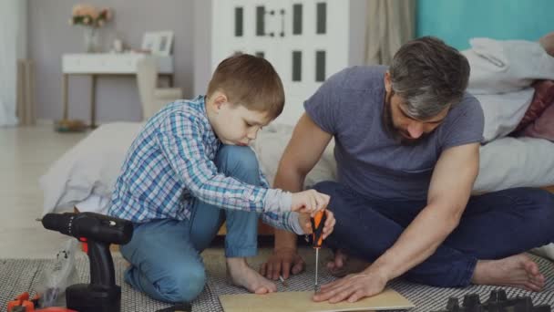 El niño serio se concentra en poner tornillo en pedazos de madera con destornillador mientras su padre lo ayuda a sostener una hoja de madera. Concepto de infancia y construcción . — Vídeos de Stock