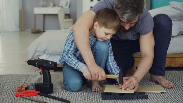 Отец-бородач учит своего сына, как использовать молоток, вбивающий гвоздь в кусок дерева, сидя дома на полу. Видны инструменты, инструменты и мебель . — стоковое видео