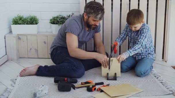 Концентрированный ребенок распиливает фанерный лист ручной пилой, а его отец держит дрова руками и помогает сыну учить его. Обмен опытом . — стоковое видео