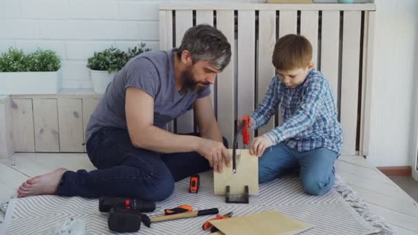 El hijo provechoso está aserrando la hoja de madera con la sierra de mano mientras que su padre está midiendo el corte con el carrete de medida y hablando con su niño. Construcción, familia unida y concepto de infancia . — Vídeos de Stock