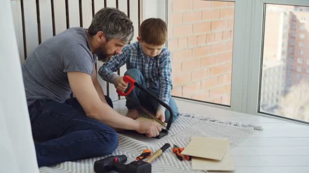 Anak laki-laki kecil difokuskan pada menggergaji sepotong kayu dengan tangan melihat dengan ayahnya membantu dan mengajarinya. Keluarga United, pekerjaan konstruksi dan konsep anak-anak . — Stok Video