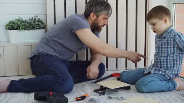 Pai amigável e filho pequeno estão conversando e rindo ao fazer o trabalho de construção sentado no chão em casa. Conversação, instrumentos e conceito de paternidade . — Vídeo de Stock