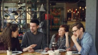 Neşeli genç arkadaş konuşurken ve paylaşım haber modern kafede masada otururken el hareketi. Büyük pizza, bardak ve tabaklar, masa ve sandalyeler görülebilir.