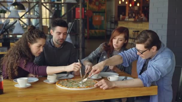 Les jeunes gens heureux coupent de grandes pizzas avec des couteaux, parlent et rient tout en dînant dans un café confortable. Style de vie moderne, manger dehors et concept d'amitié . — Video