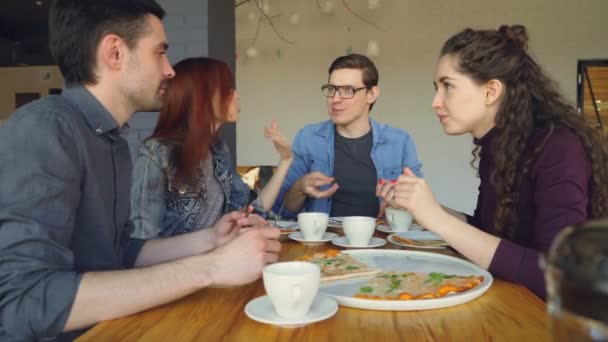 Šťastní studenti sdílejí novinky, gestikuloval a smáli se při jídle lahodné pizzy v pěkné útulné kavárně. Chutné jídlo, veselé lidi a jídelní z koncepce. — Stock video