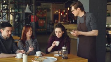 Dost gülümseyen garson önlük sipariş deftere yazarken çekici arkadaş kafede yiyecek ve içecekler sipariş ettiğiniz. Genç insanlar, dostluk ve gıda kavramı.