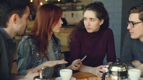 Des jeunes hommes et femmes joyeux et proches utilisent leur smartphone et parlent tout en déjeunant dans un bon café. Salle à manger, technologie moderne et concept d'amitié . — Video